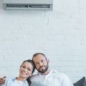 Couple relaxing indoors with a wall-mounted air conditioner, celebrating comfort and the importance of HVAC systems on National AC Day.