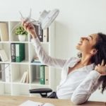 Woman in an office happily holding a pair of shoes, surrounded by a desk, chair, bookshelves, and a small plant.