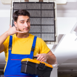 Young man in work attire gesturing with a phone, holding a toolbox near an air conditioning unit for Easco HVAC services.
