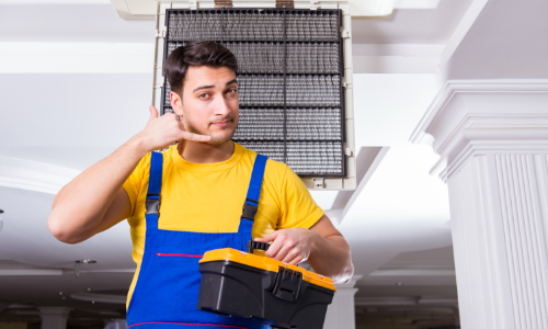 HVAC technician in blue overalls holding a toolbox, signaling a phone call with an air conditioning unit in the background, emphasizing professional HVAC maintenance and repair services.