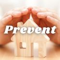 Hands covering a wooden house model with "Prevent" text, emphasizing home safety in HVAC services.