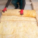 Individual wearing gloves installs yellow fiberglass insulation over wooden flooring for HVAC home improvement.