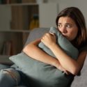 Young woman sitting on a couch in a living room, showing signs of distress amidst a cozy indoor setting.
