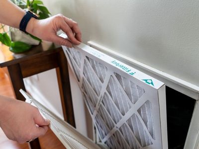 Individual replacing a clean Filtrete air filter near an air intake vent in an organized living space by Easco HVAC services.