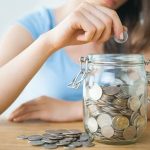 Young person placing a coin into a clear jar filled with coins for savings.