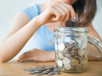 Person placing a coin into a glass jar filled with coins, symbolizing savings and financial efficiency, relevant to geothermal system investment benefits.