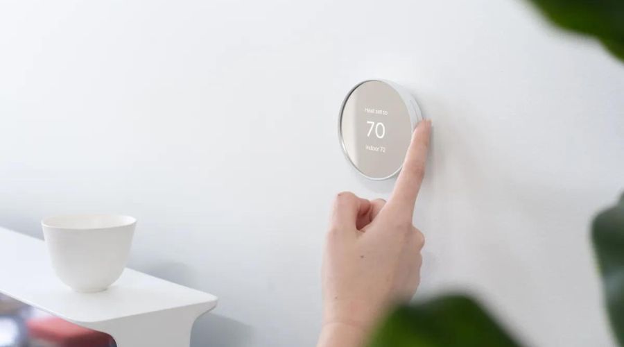 Smart thermostat on wall being adjusted, displaying temperature settings, with a minimalist design and a nearby white bowl, emphasizing energy efficiency and modern home technology.