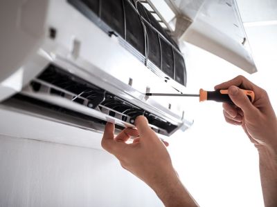 Person performing maintenance on a wall-mounted air conditioning unit, using a screwdriver to adjust the front panel.