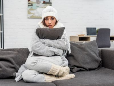 Person sitting on a couch, bundled in warm clothing, hugging a pillow, looking uncomfortable in a cold indoor setting, highlighting issues of inadequate heating and comfort related to HVAC systems.
