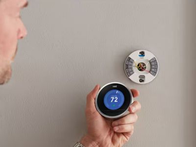 Person installing a smart thermostat, displaying temperature settings, next to an existing thermostat on a wall, illustrating HVAC efficiency and home automation.