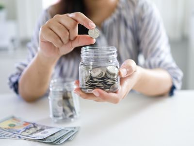 Person placing a coin into a glass jar filled with coins, with dollar bills visible on the table, illustrating savings and financial management concepts.
