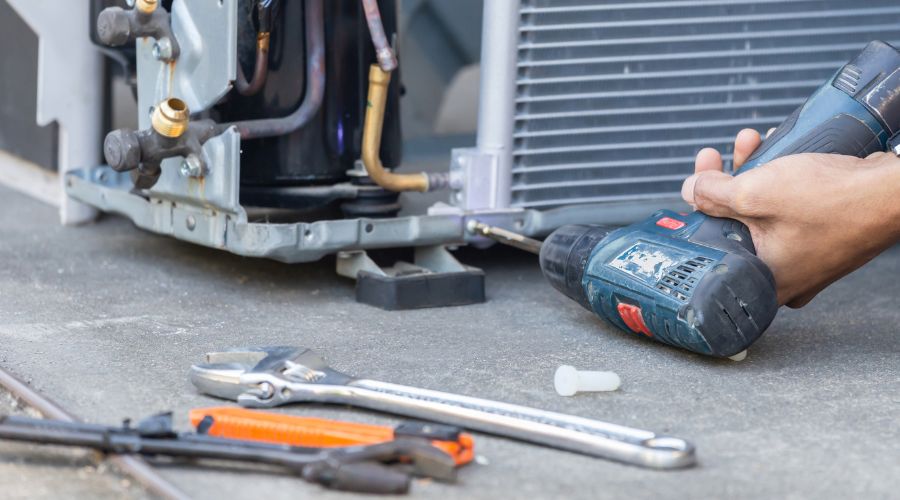 Air conditioning technician using a power drill to repair an AC unit, surrounded by various tools, illustrating the importance of professional HVAC services for efficient air conditioning maintenance.