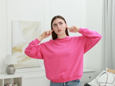 Woman in a pink sweatshirt covering her ears, reacting to loud sounds from an air conditioning unit, illustrating the need for AC repair.
